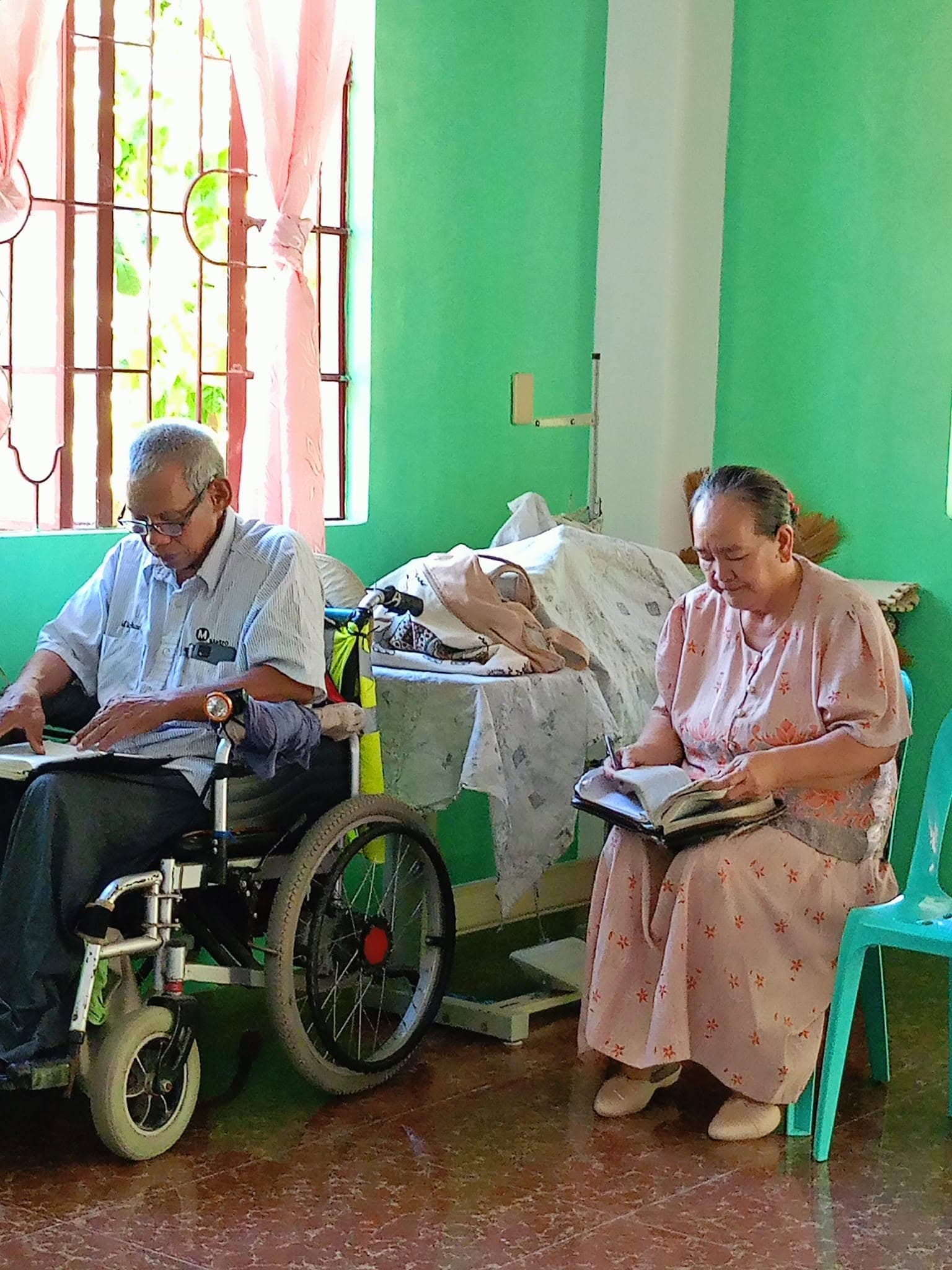 An elderly couple seated side by side, heads bowed over open Bibles. One of them holds a pen, taking notes while reading.